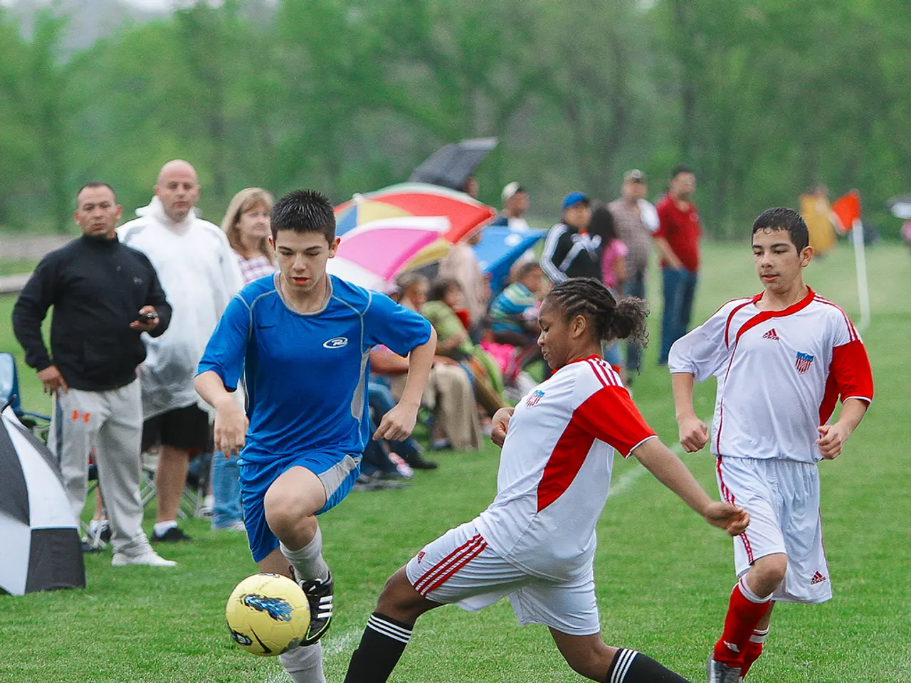 There are three persons playing football. Behind them there are group of people standing and...