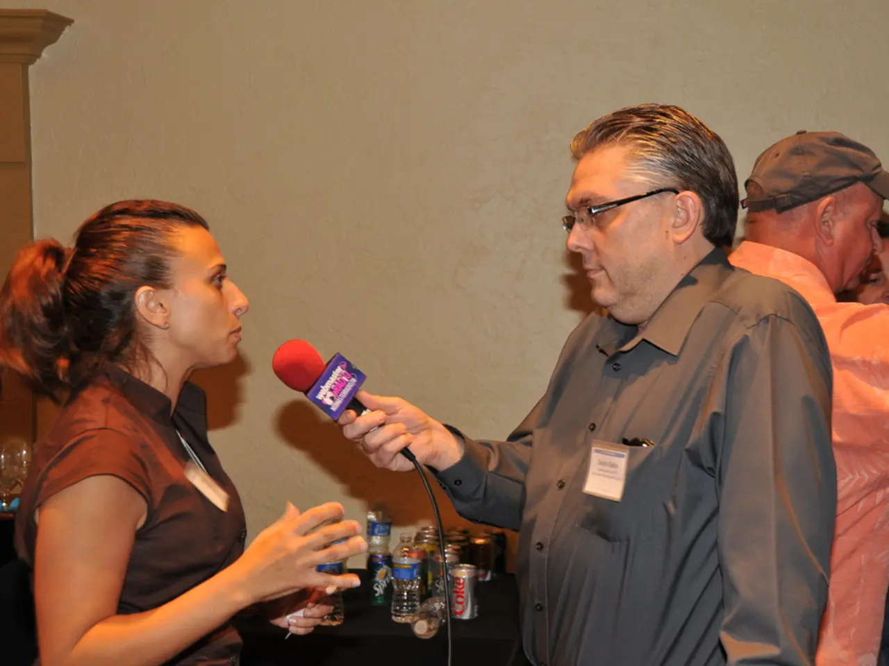 In this picture a guy is holding a mic in his hand and interviewing a lady. To the right side of...
