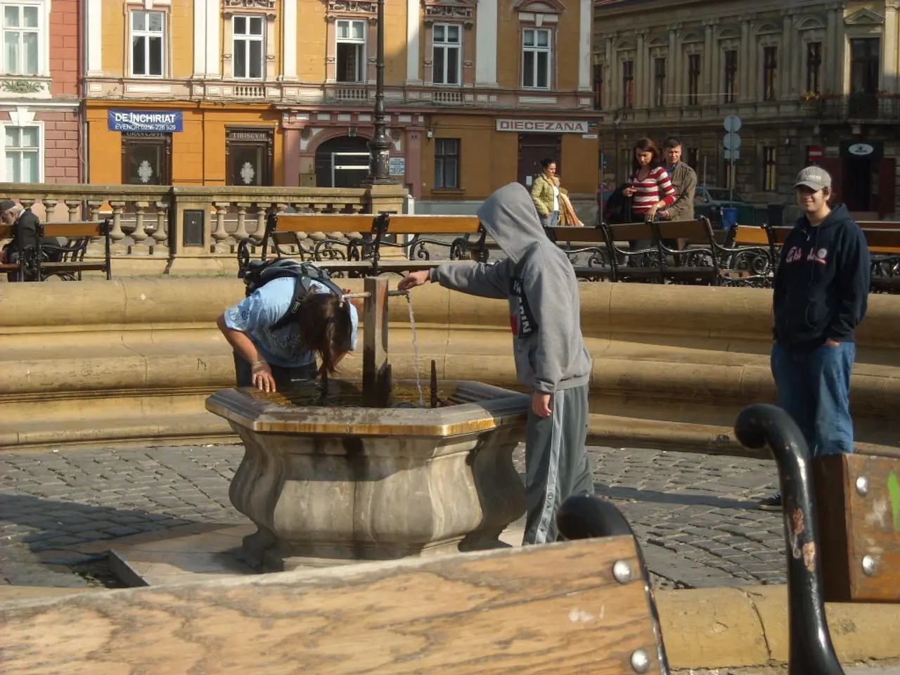 In the center of the image there is a fountain. Beside the fountain there are people. Behind the...