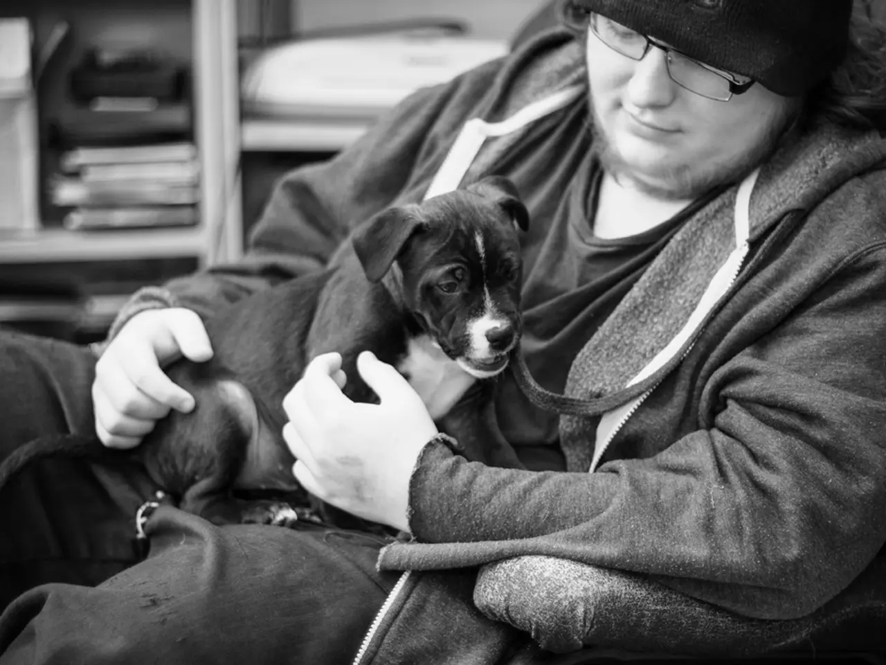 There is a man who is wearing spectacles and monkey cap pampering a puppy by placing it on his...