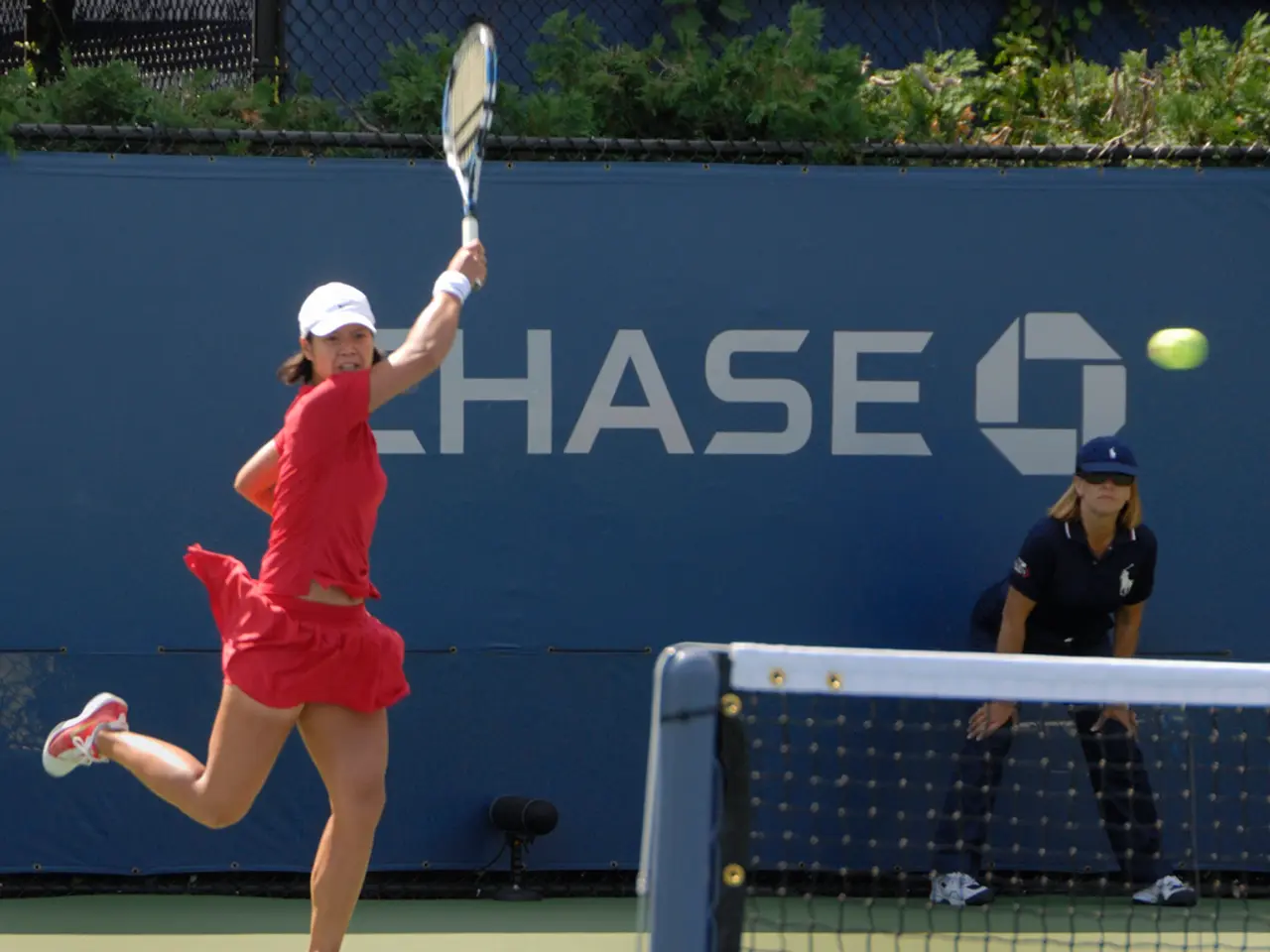 The woman in red T-shirt is holding a racket in her hand and I think she is playing tennis. In the...