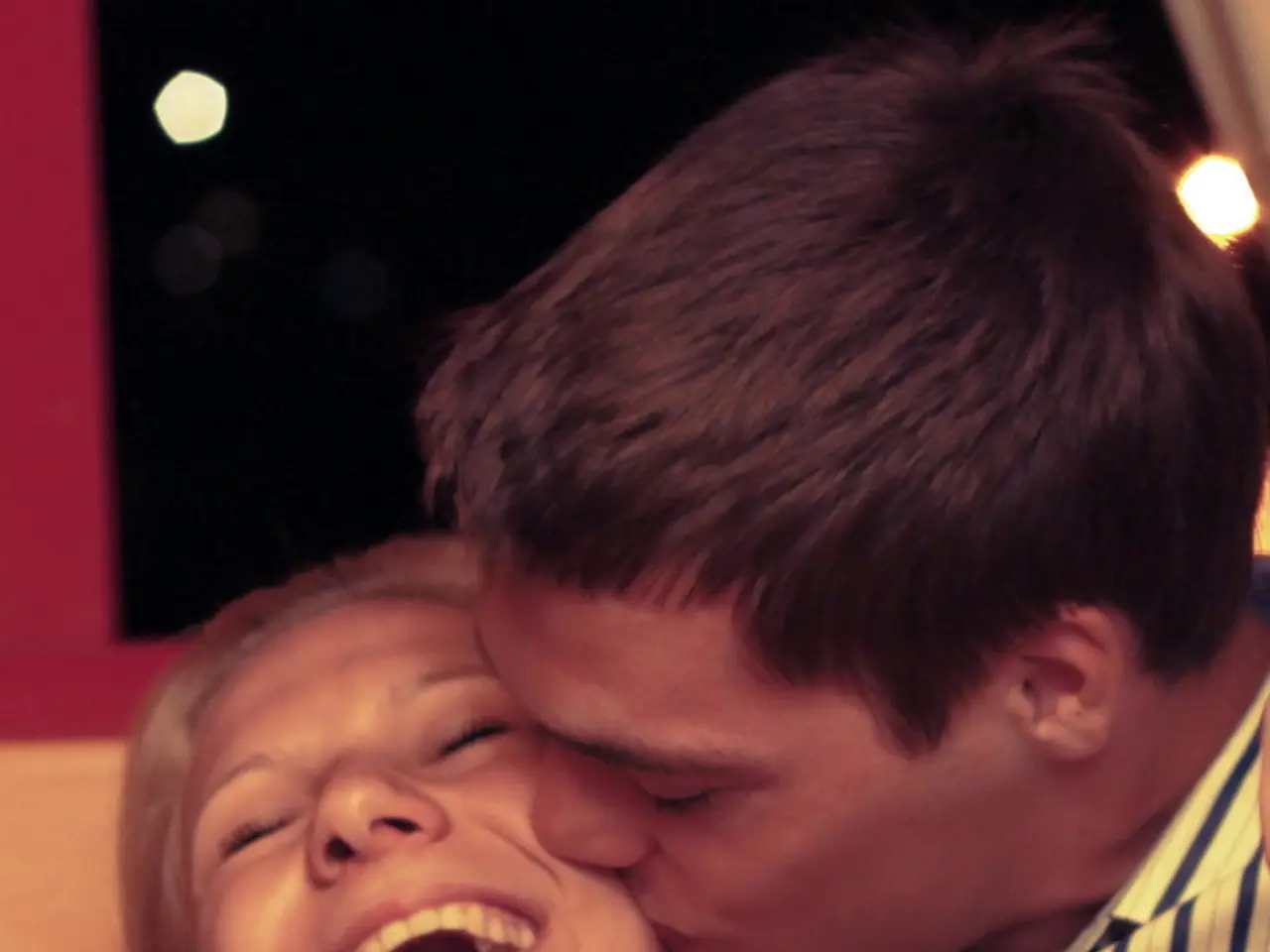 In this picture we can see a man kissing a woman and she is smiling and in the background we can...