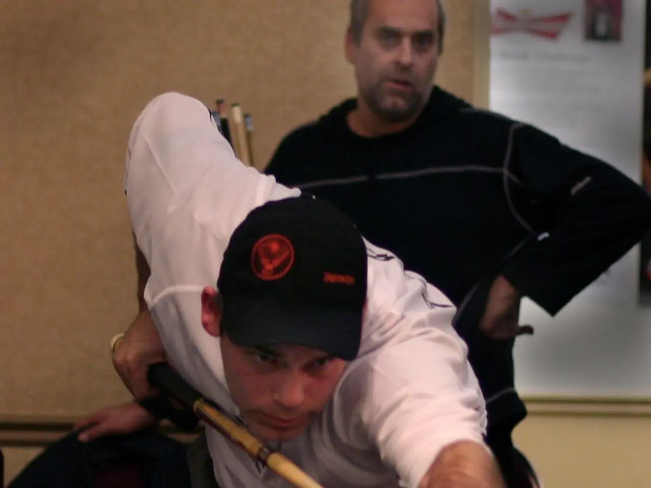 In this image, man in white t-shirt who is wearing black cap is playing snooker pool game. Behind...