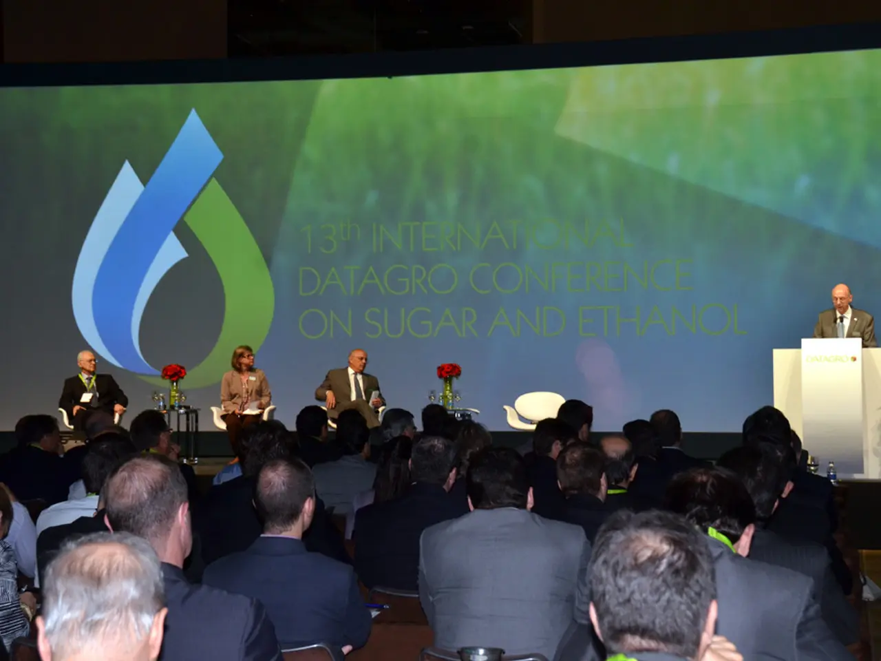 In this picture we can observe some people sitting in the chairs in front of a stage. We can...