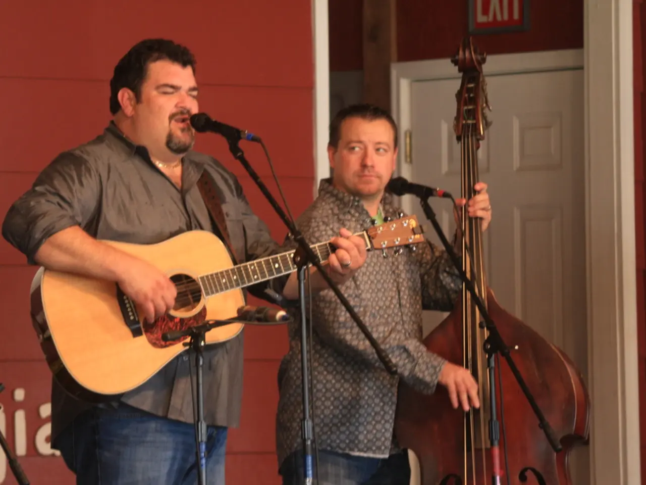 A man singing with mic in front of him playing a guitar. Another man beside him is playing cello.