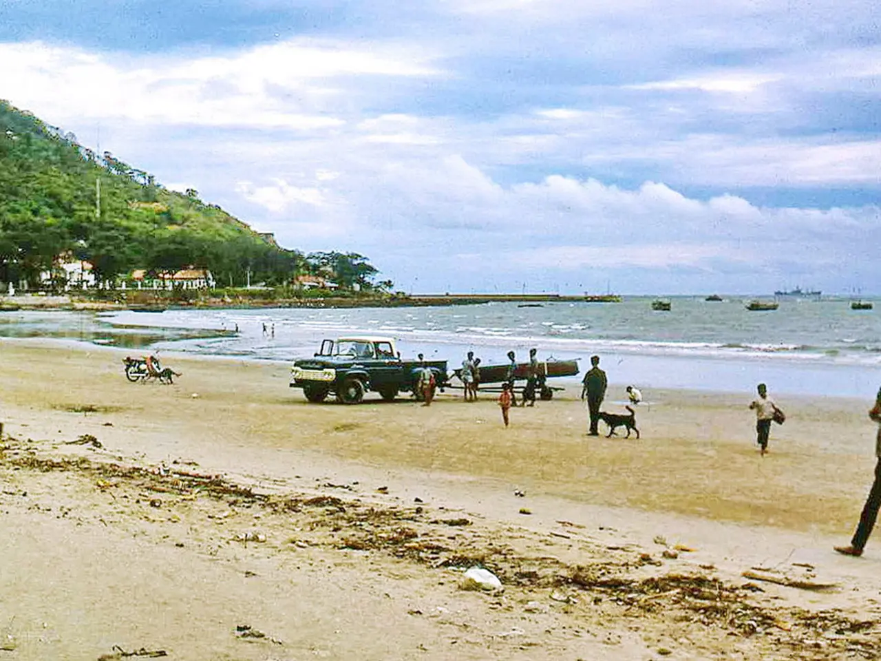 In this image there are people walking on a beach and there is a vehicle, in the background there...