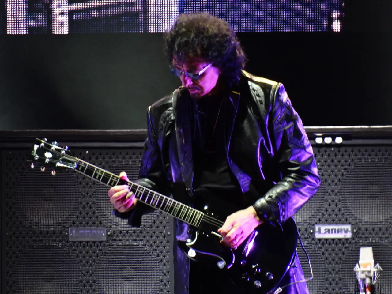 In this image I see a man who is wearing a jacket and is holding the guitar. In the background I...