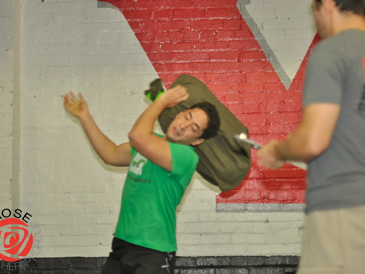 This image consists of two persons. In the front, the man wearing green T-shirt is holding a bag....