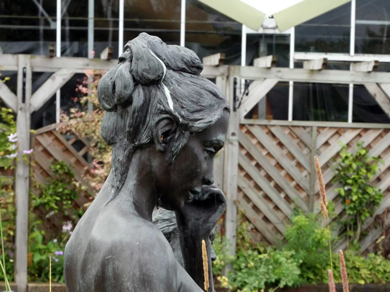 This picture shows statue of a woman. We see plants and a wooden fence.