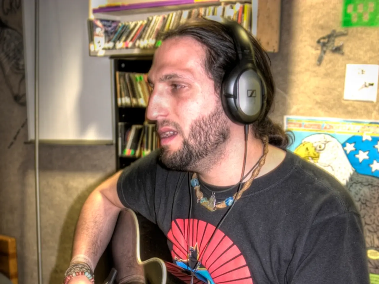 In this picture a black shirt guy is playing a guitar and wearing a headphones. In the background...