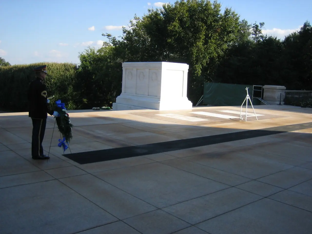 This picture shows a man standing and holding a stand with flowers to it, Man wore a cap on his...