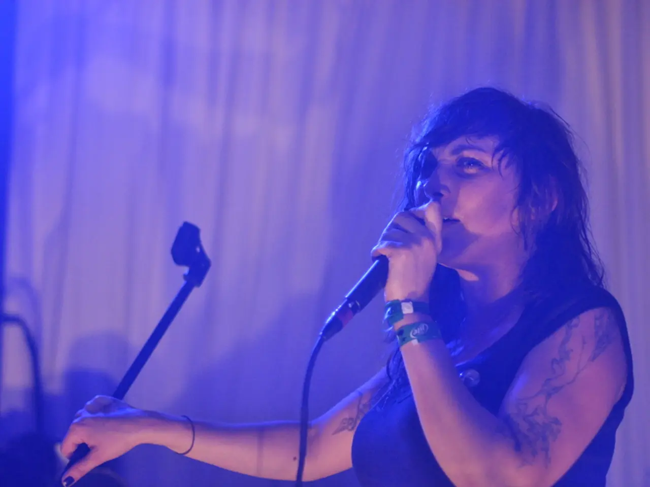 The image consists of a lady singing on mic ,she had tattoo on her hands.