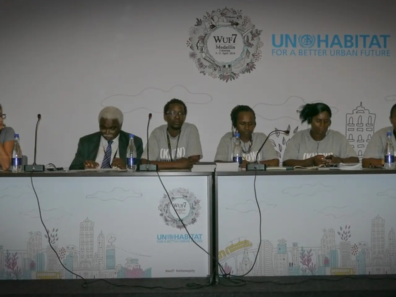 This image is clicked in a conference hall. There are six persons sitting in this image. To the...