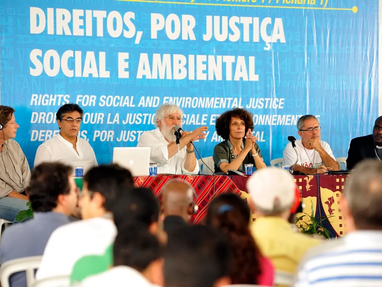 In this image there are six persons sitting on a stage, in front of them there is a table, on that...