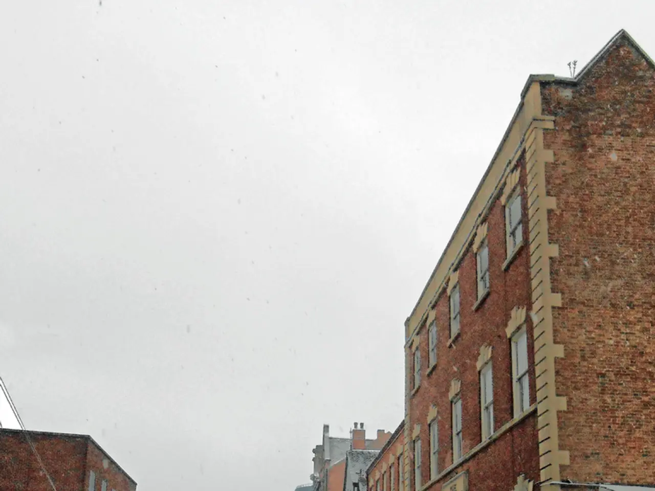 In this image there is a road in the middle. On the road there is snow. There are buildings on...