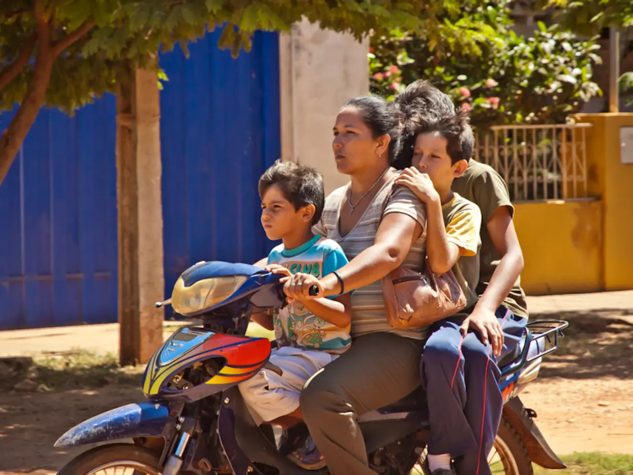 Here we can see a woman driving a motor cycle where a boy sitting in front and there are other two...