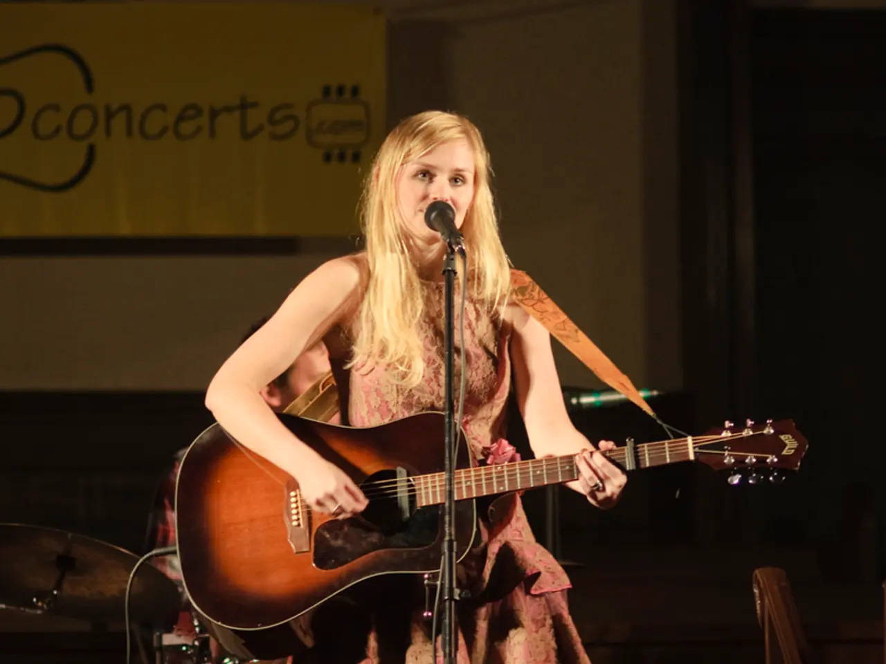 This image is clicked in a concert. In the front, there is a woman playing guitar and singing. In...