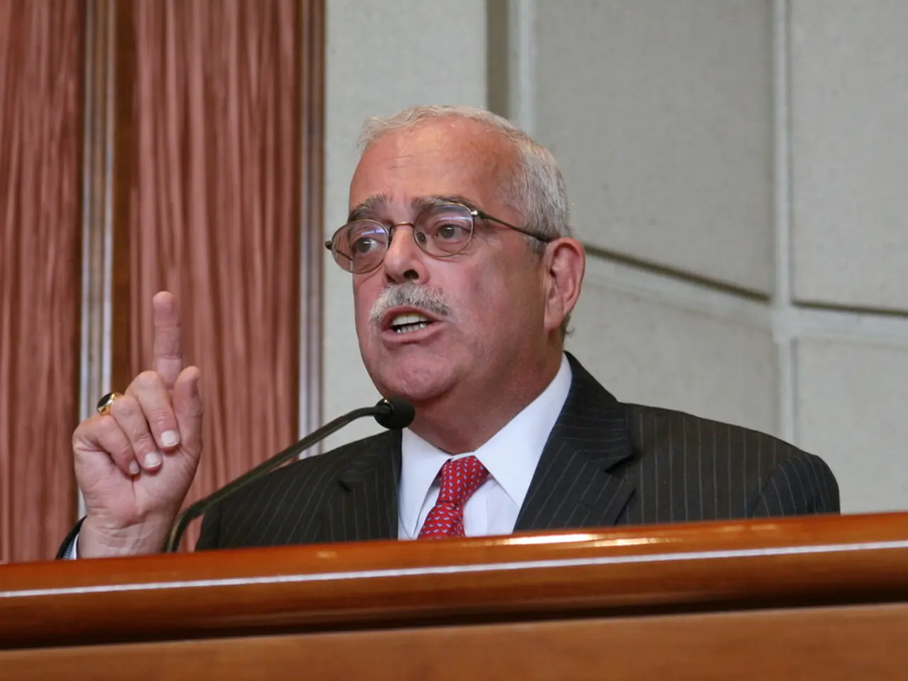 There is a person wearing specs and black suit is speaking. In front of him there is a mic and a...