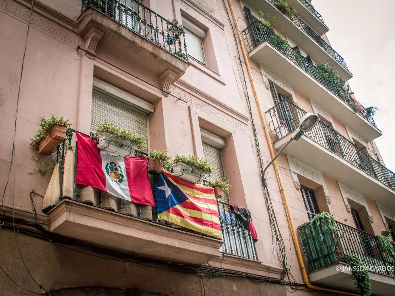 In this image there are buildings and in front of the building there are flags on the balcony and...