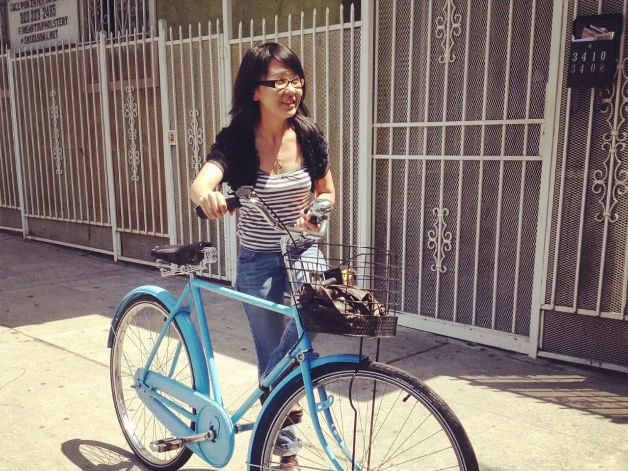 In this image in the center there is one woman who is holding a cycle and walking, and there is one...