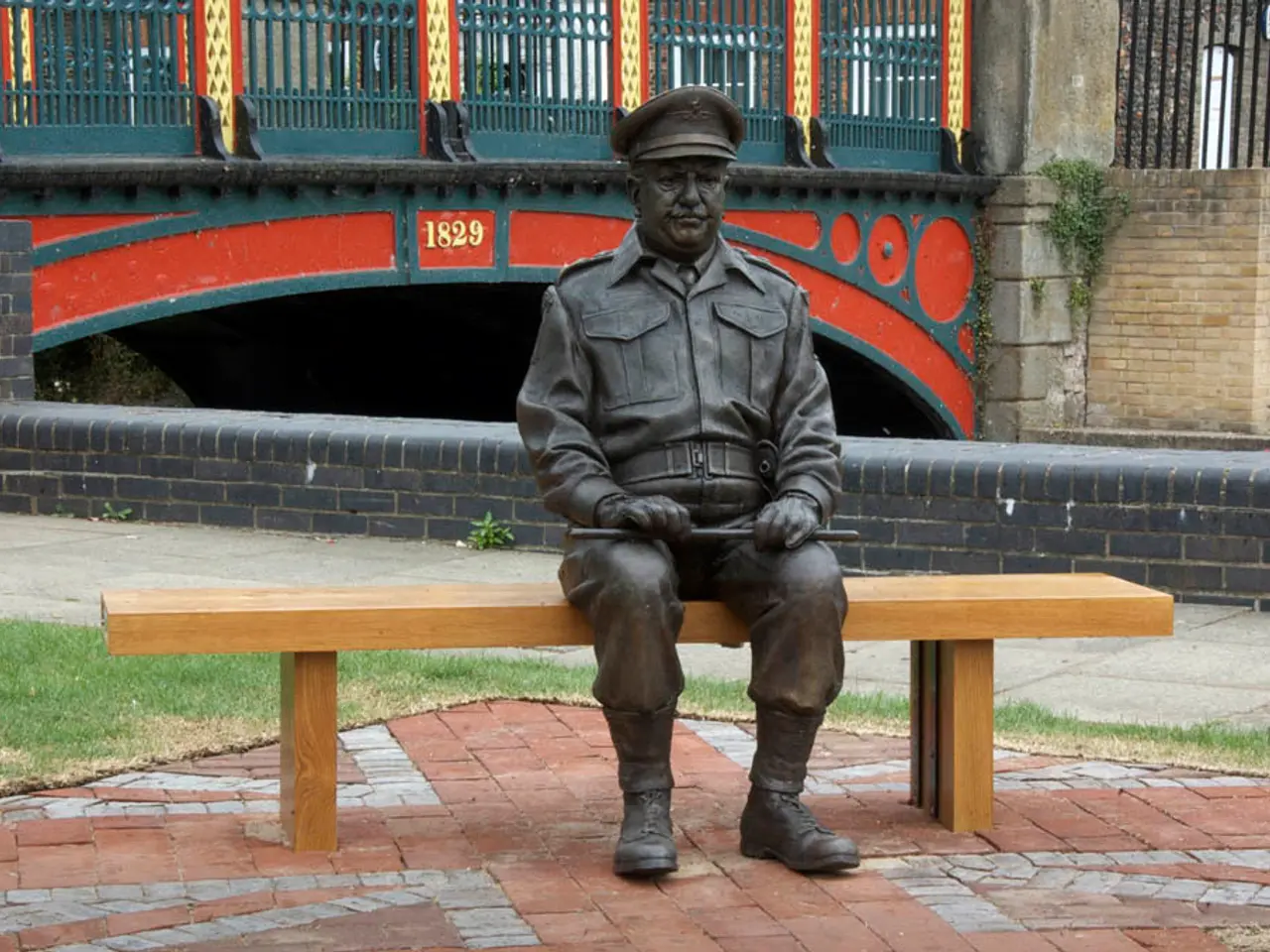 This is a iron statue of a man which is on the cardboard bench. Behind him there is a bridge like...