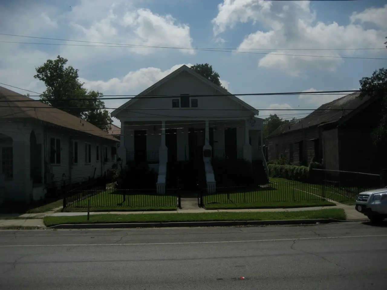 Sky is cloudy. Here we can see buildings, grass, fence, vehicle and trees.