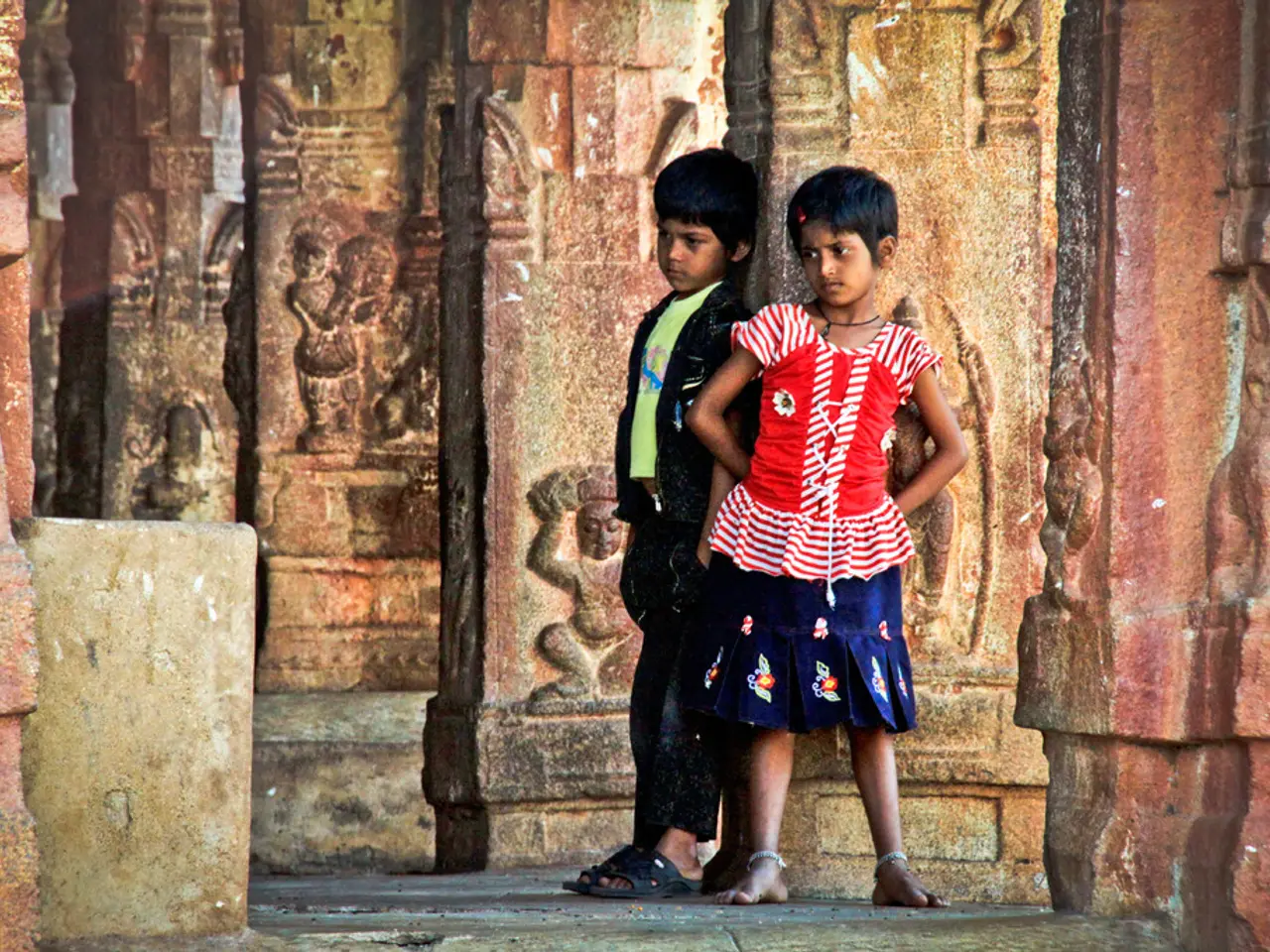 In this image two children are standing and one of the girl is wearing red color dress,and a boy is...
