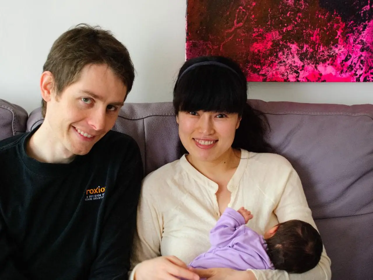 Woman in a white shirt who is holding a baby in her hand is sitting on the sofa and she is smiling....