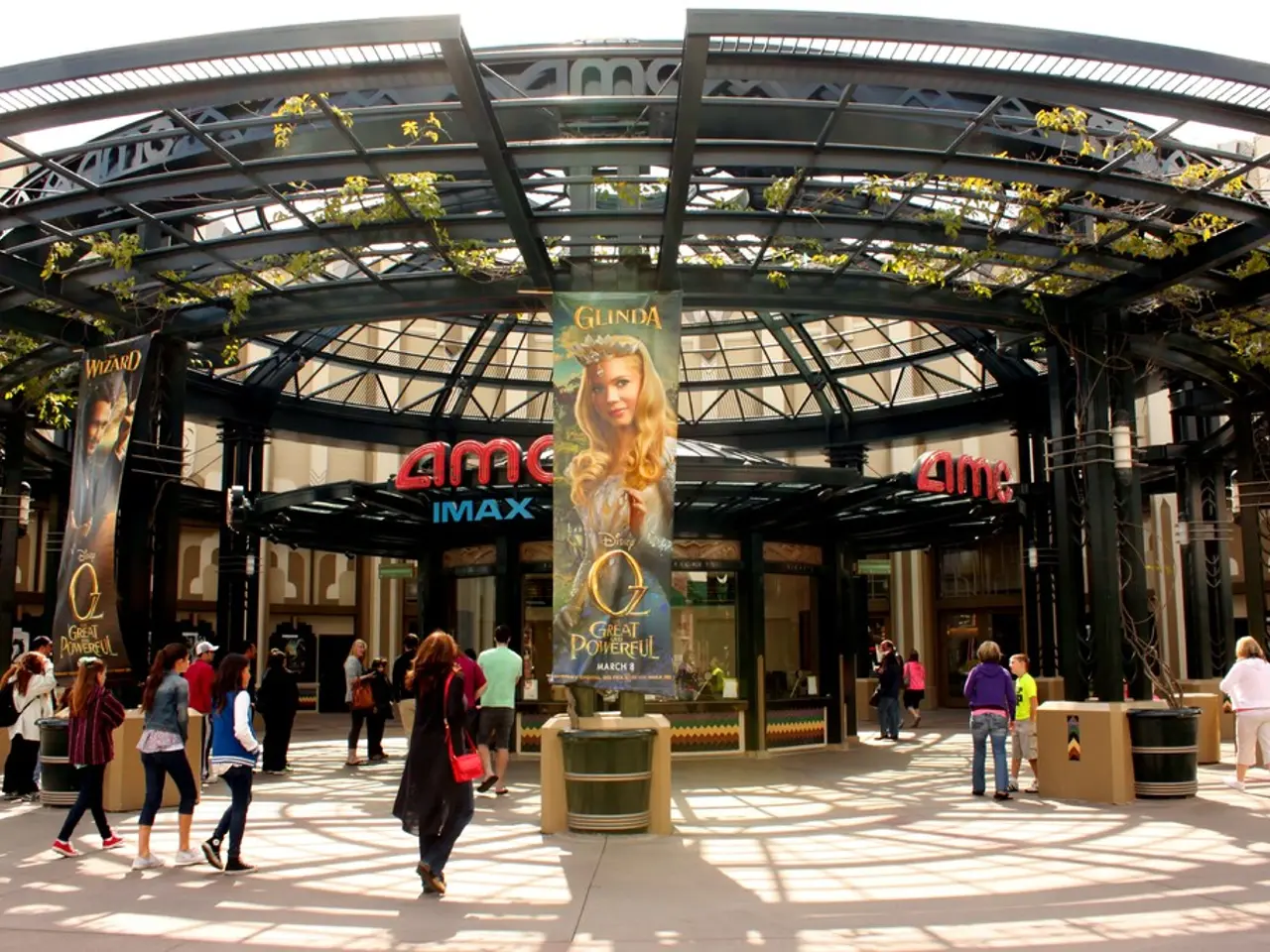 In this image there are banners. There are dustbins. There are people walking. In the background of...