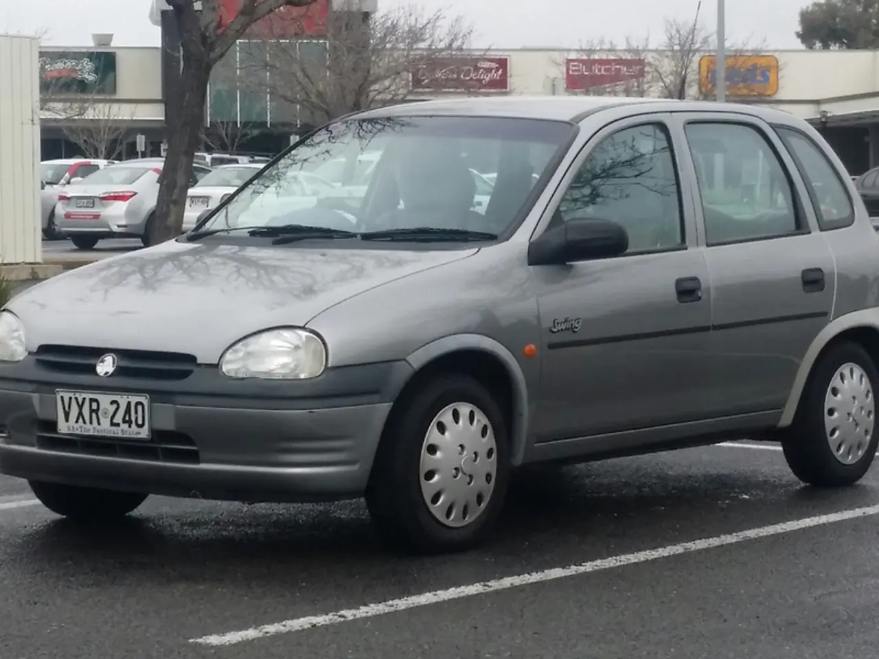 There are vehicles on the road and there is a bin. Here we can see trees, plants, boards, poles,...