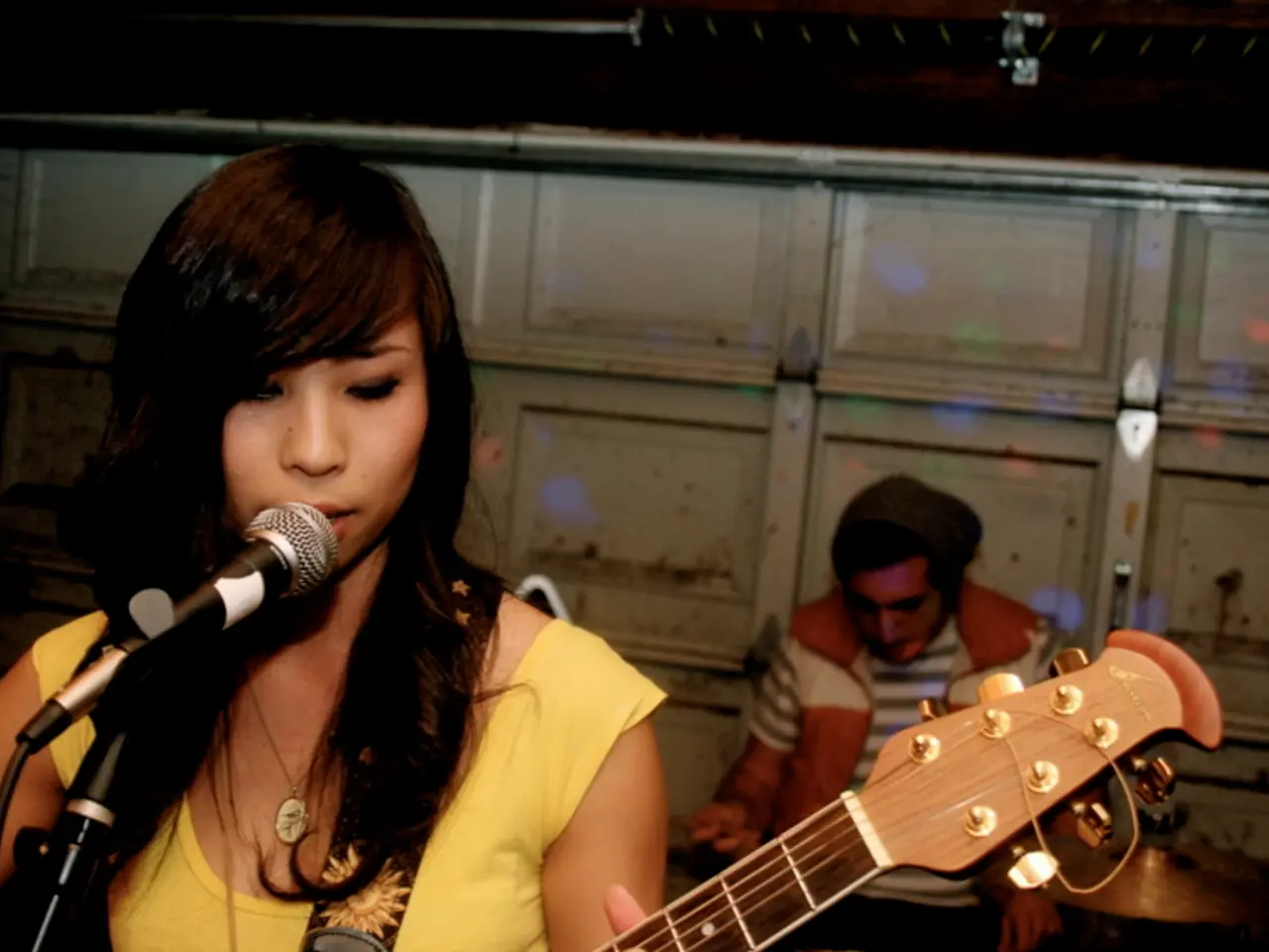 The picture is is of a live performance. In the foreground a woman is playing guitar and singing....