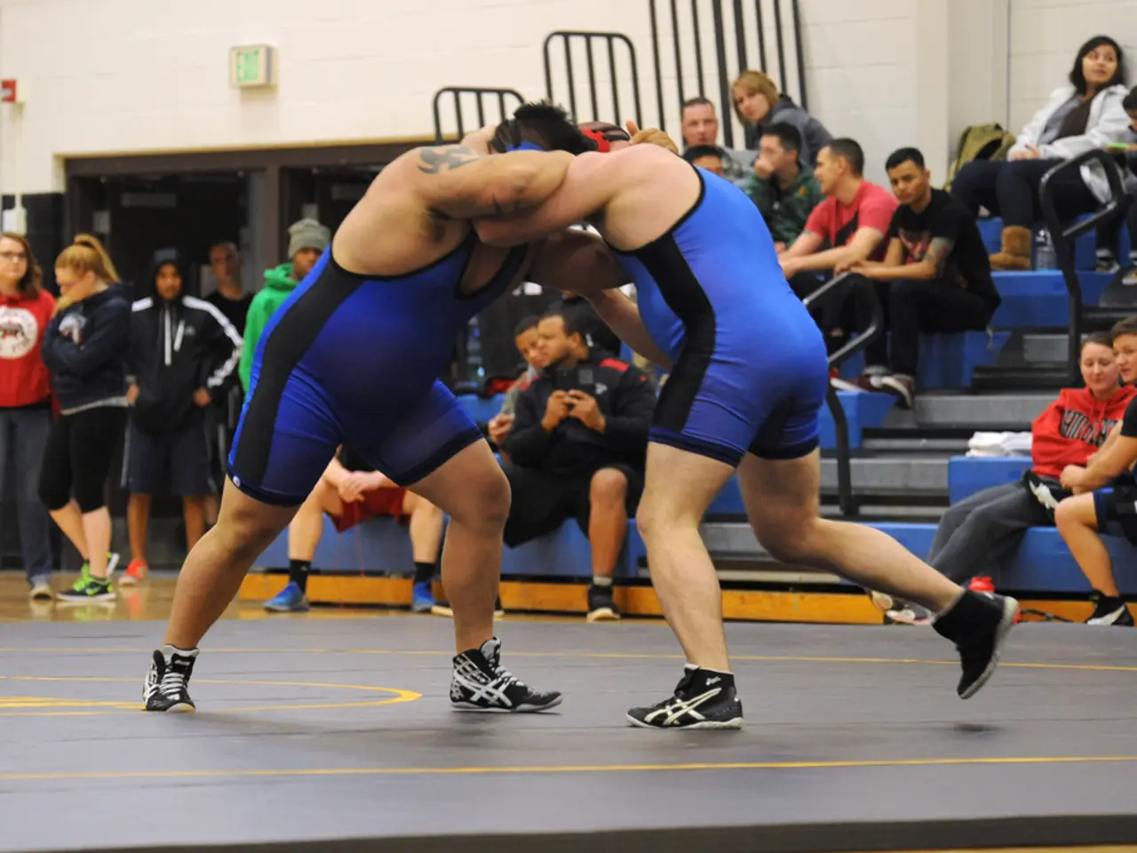 In the image two persons are doing wrestling. Behind them few people are sitting and standing and...