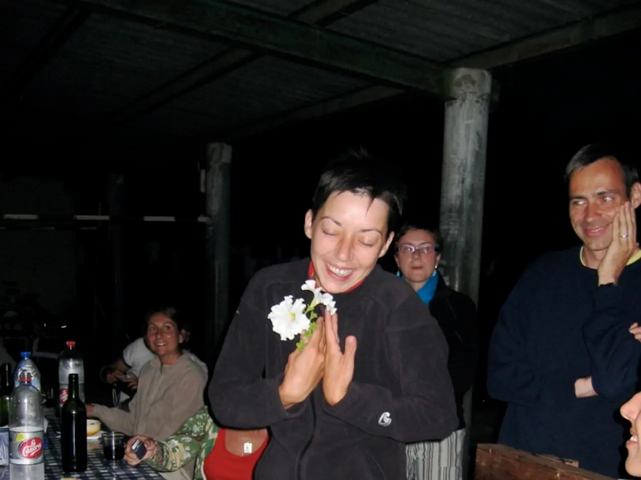 A woman is standing and also laughing. Left side there are plates,food,glasses,bottles on the...