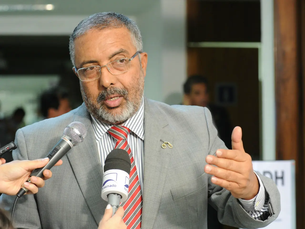 In this image there is a man, he is wearing suit and talking to journalists who are holding mics in...