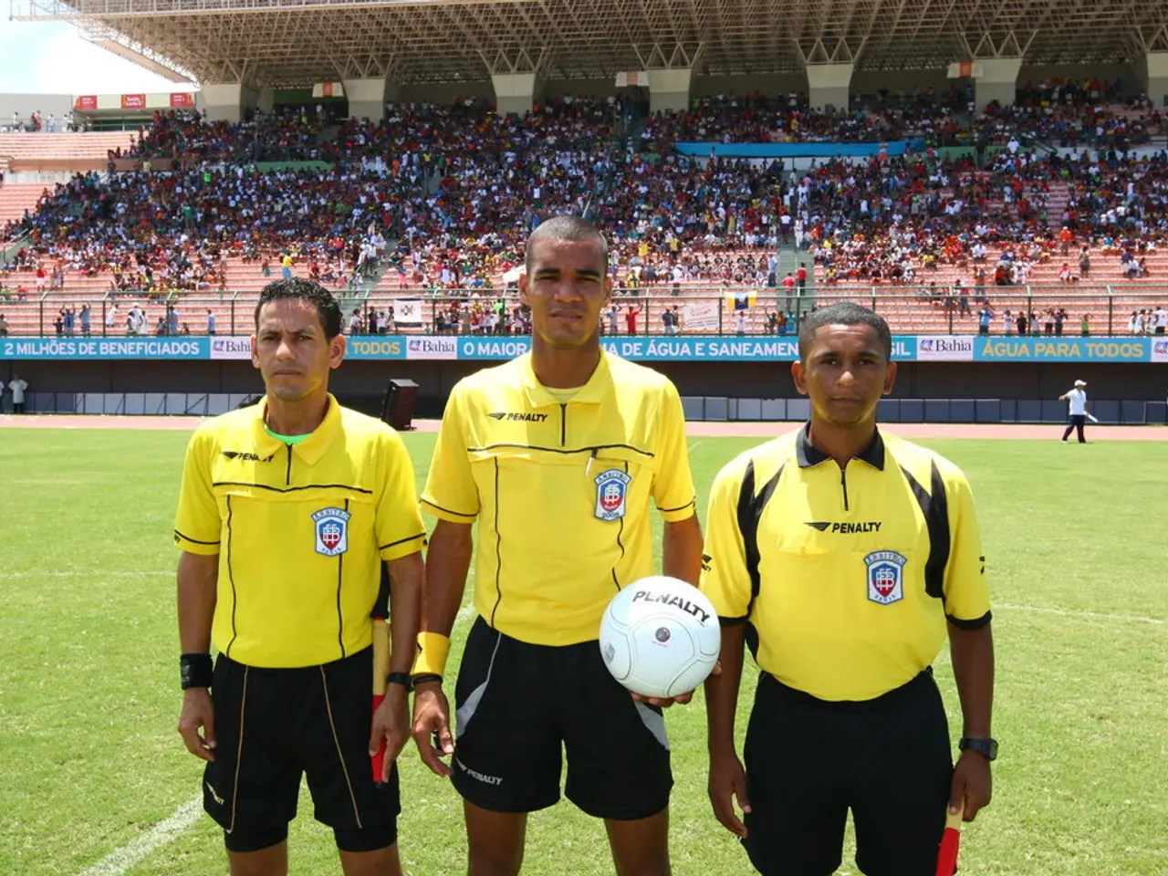 This picture is taken in a stadium. In the foreground there are three people standing. The right,...