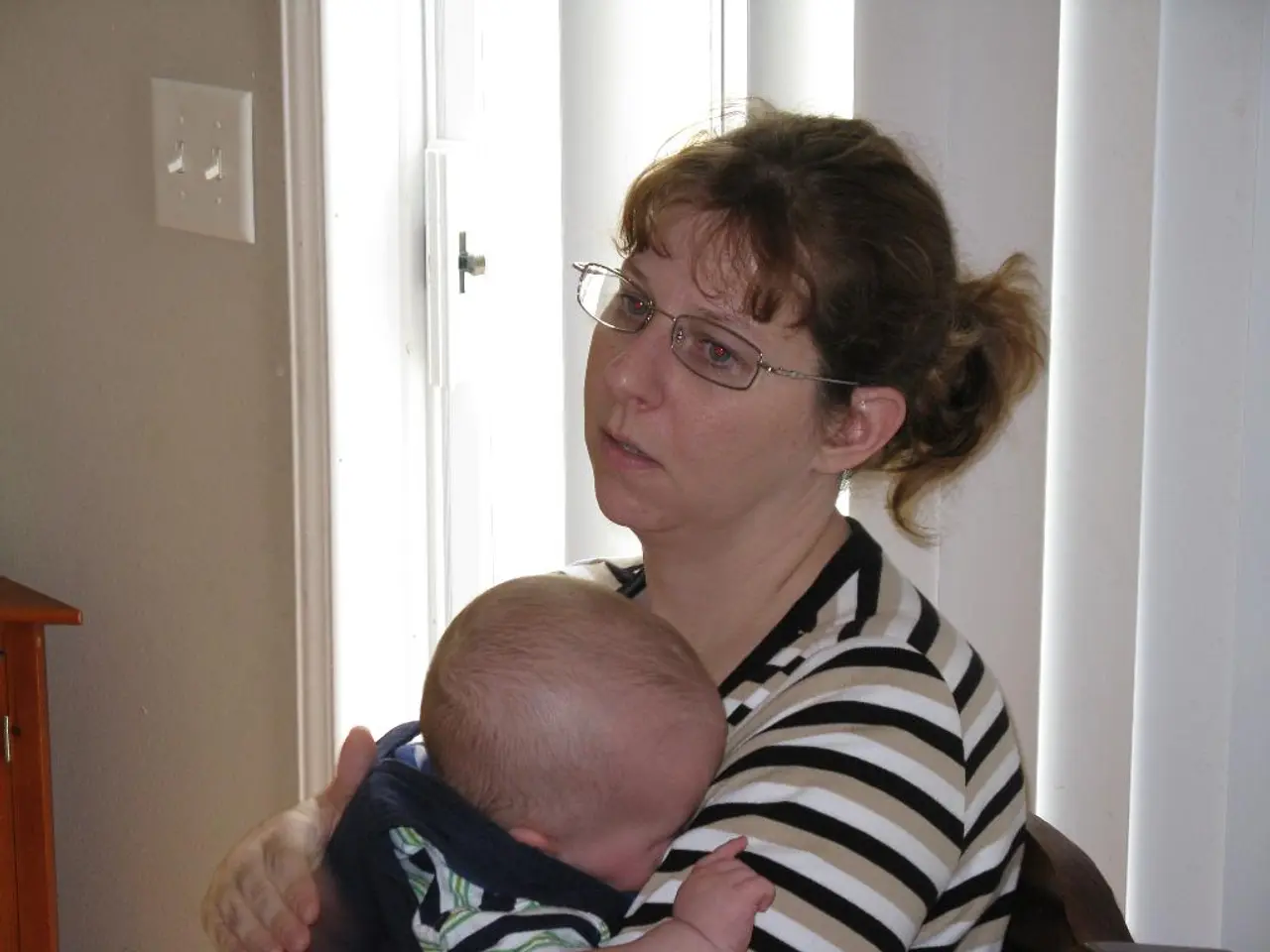 In this image there is a woman sitting on the chair. She is holding an infant in her hand. Behind...