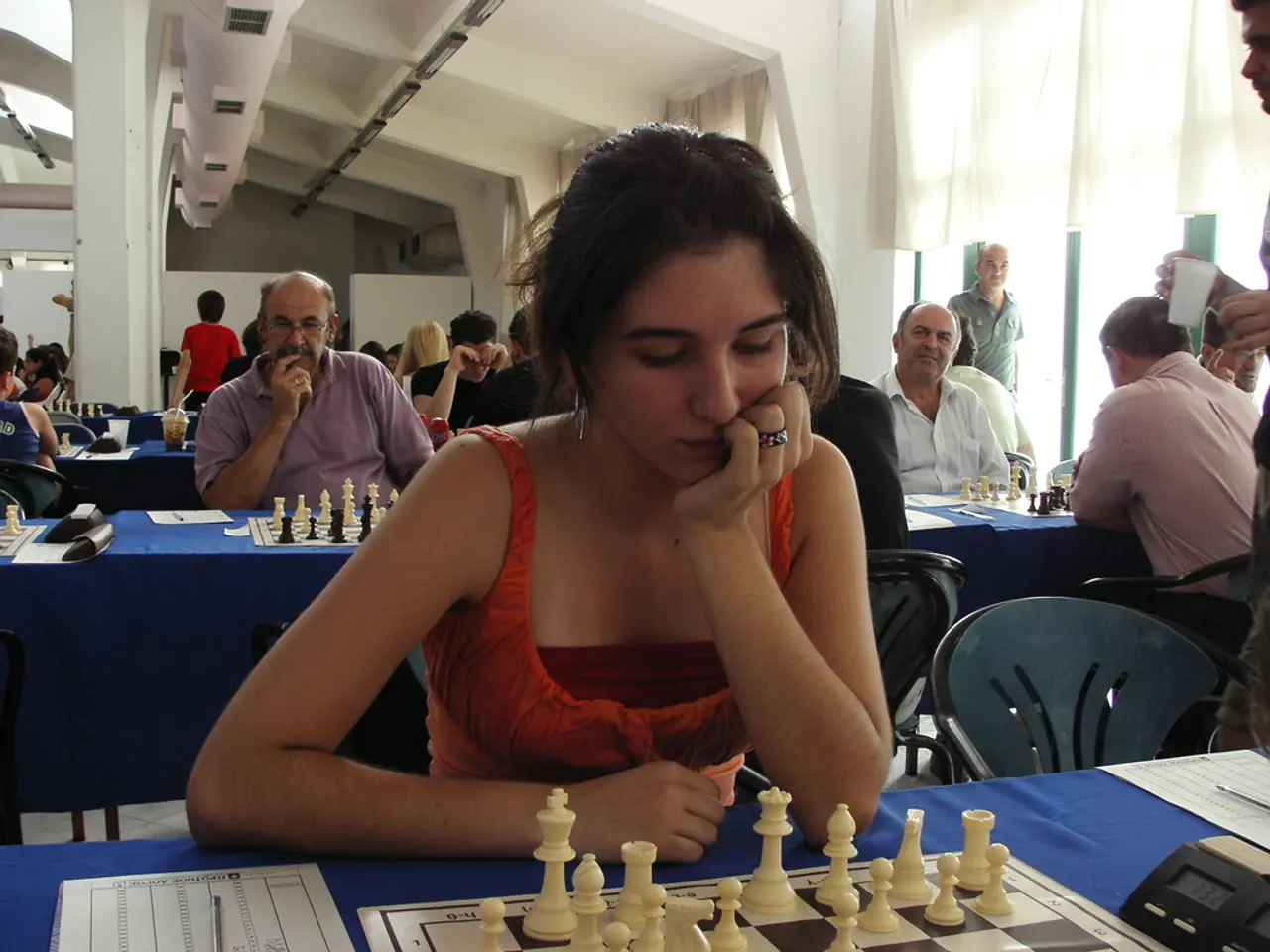 The women wearing a red dress is playing chess which is placed on a table with a blue mat on it and...