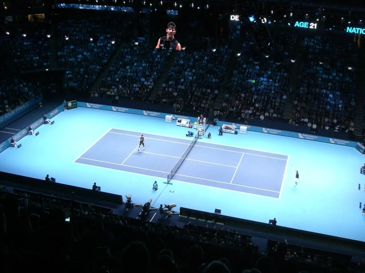 There is a tennis court. Two persons are playing. On the sides of the court there are banners....