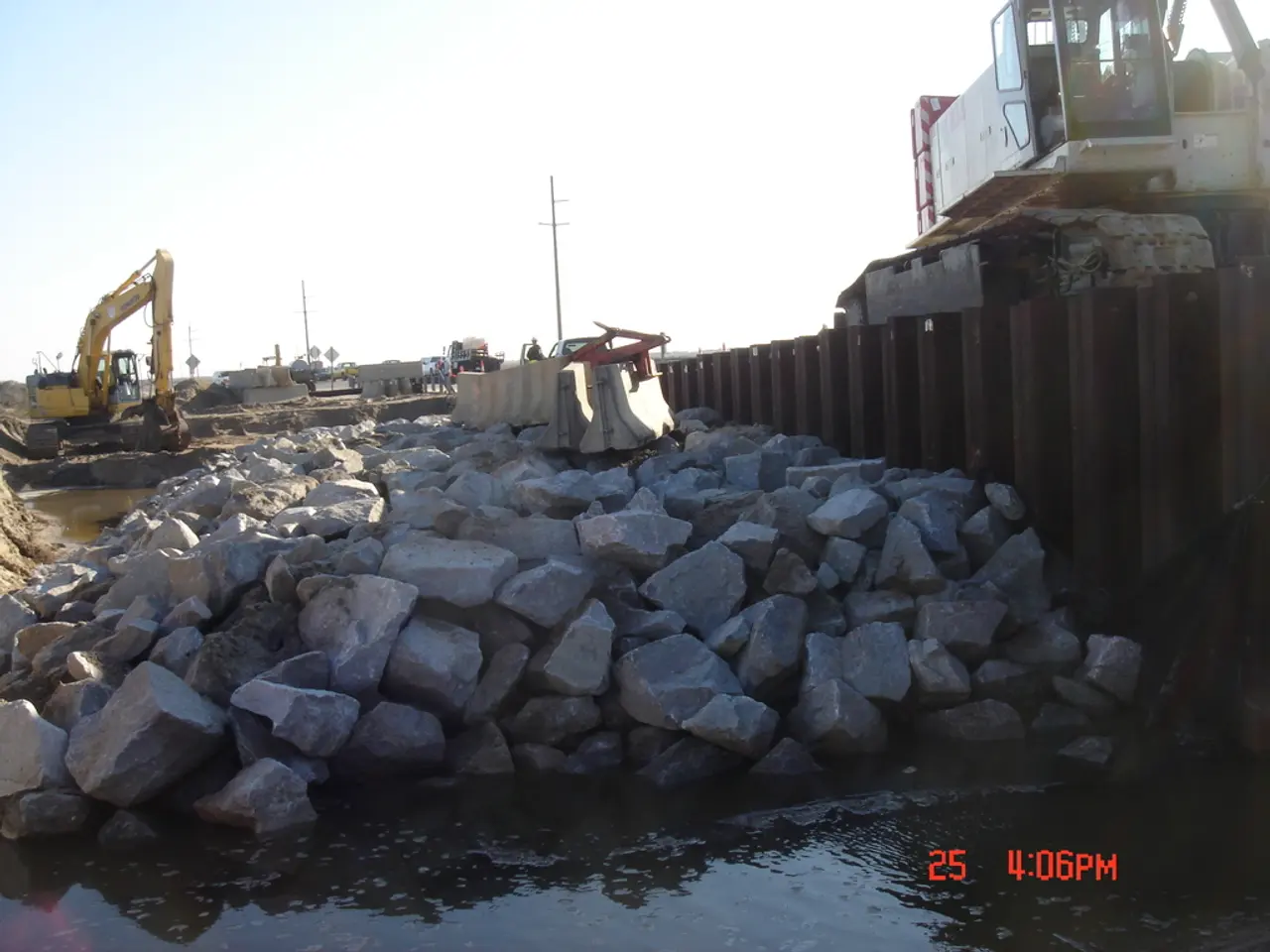 In this image we can see rocks, water, iron grill, sand, excavators, poles and sky.