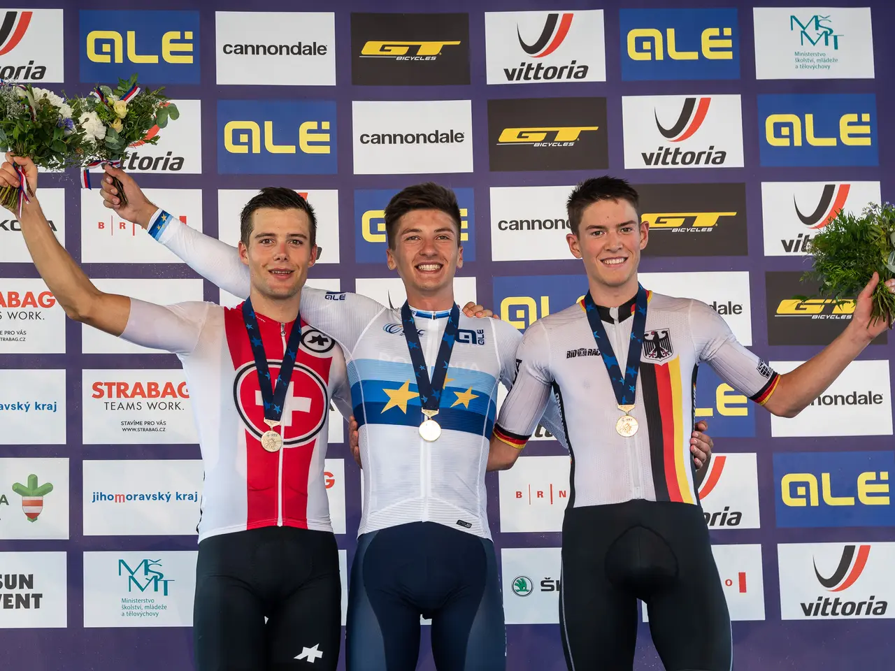 The image shows three men standing on top of a podium, each wearing a medal and holding a bouquet...