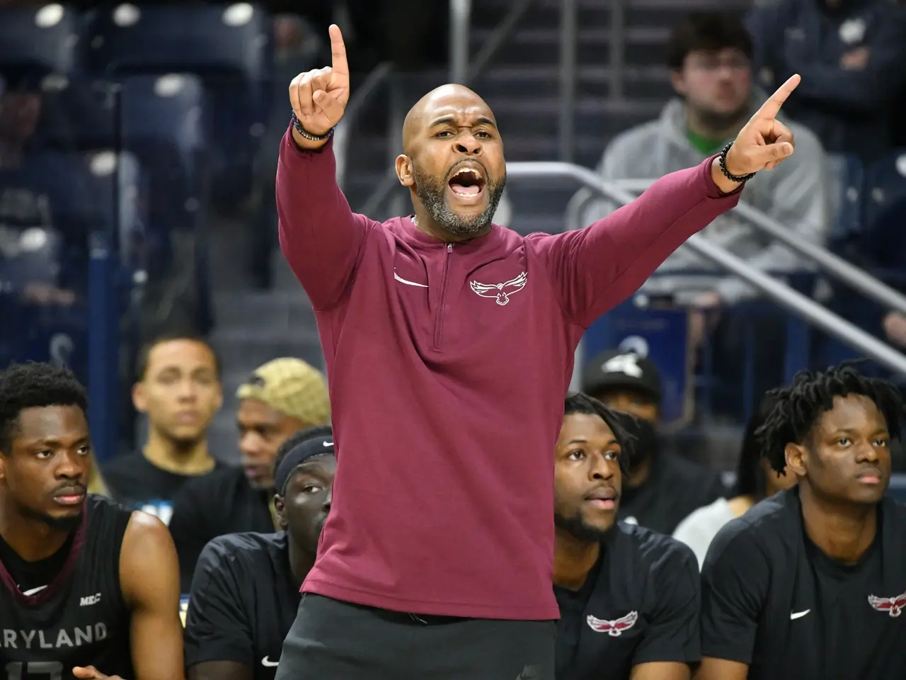 The image shows a man in a maroon shirt and black shorts on the sidelines of a basketball court,...