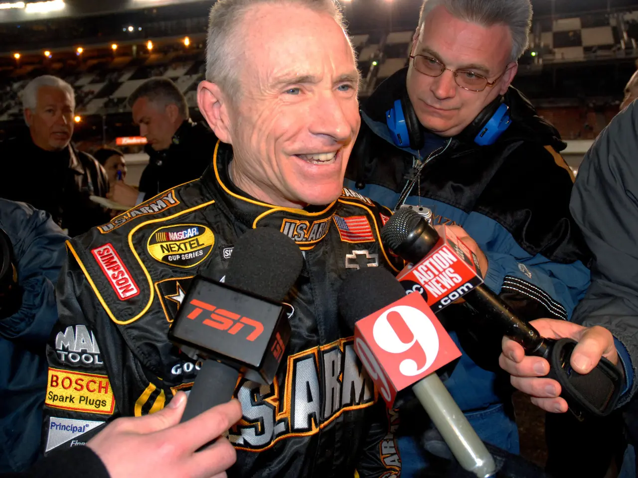 The image shows a man in a black jacket talking to reporters in front of a stadium filled with...
