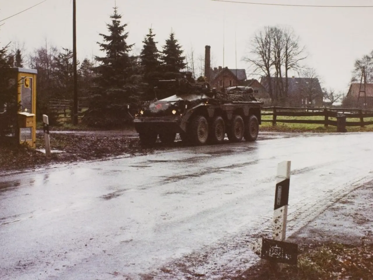 The image shows an armored vehicle driving down a snowy road, surrounded by a wooden fence, grass,...
