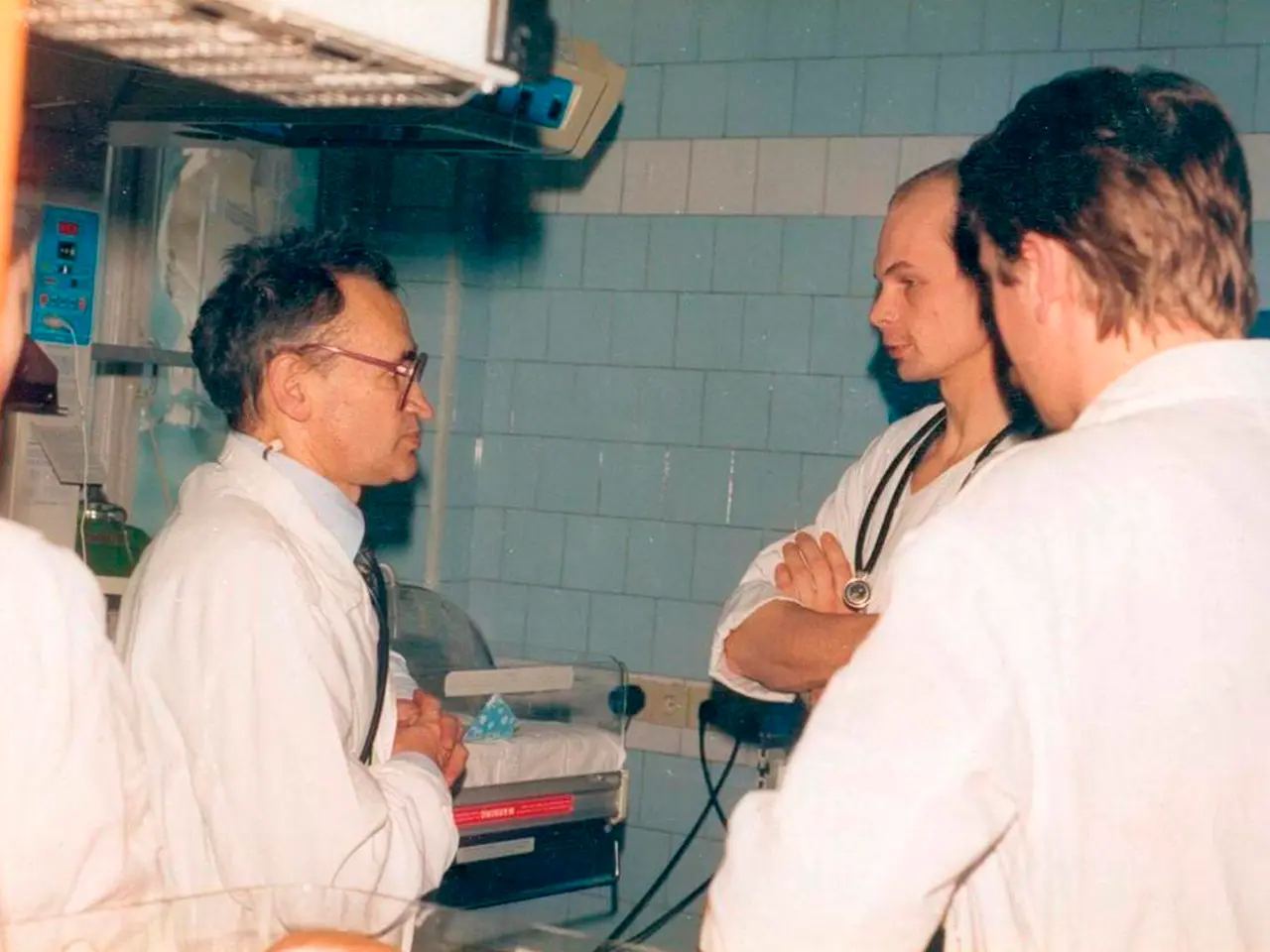 The image shows a group of three men in white lab coats standing around a hospital bed, with a wall...