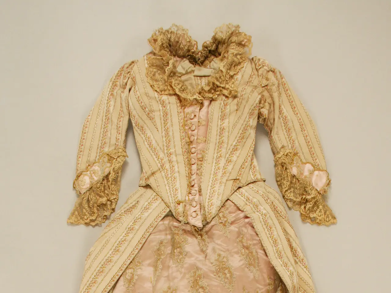 The image shows a pink and gold dress on display in a museum, with a cream-colored background. The...