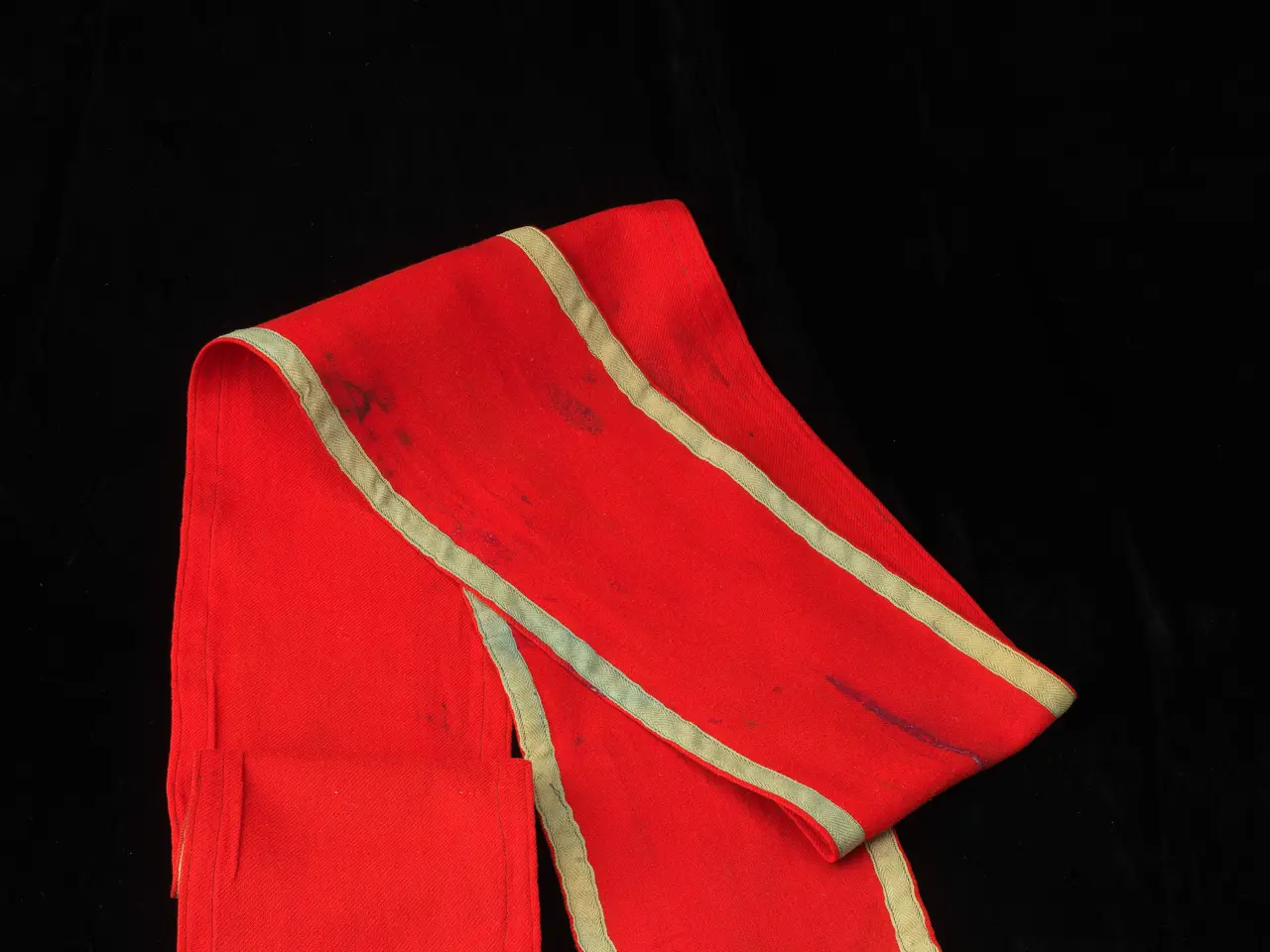 The image shows a red stole with gold trim on a black background. The stole is a deep red color...