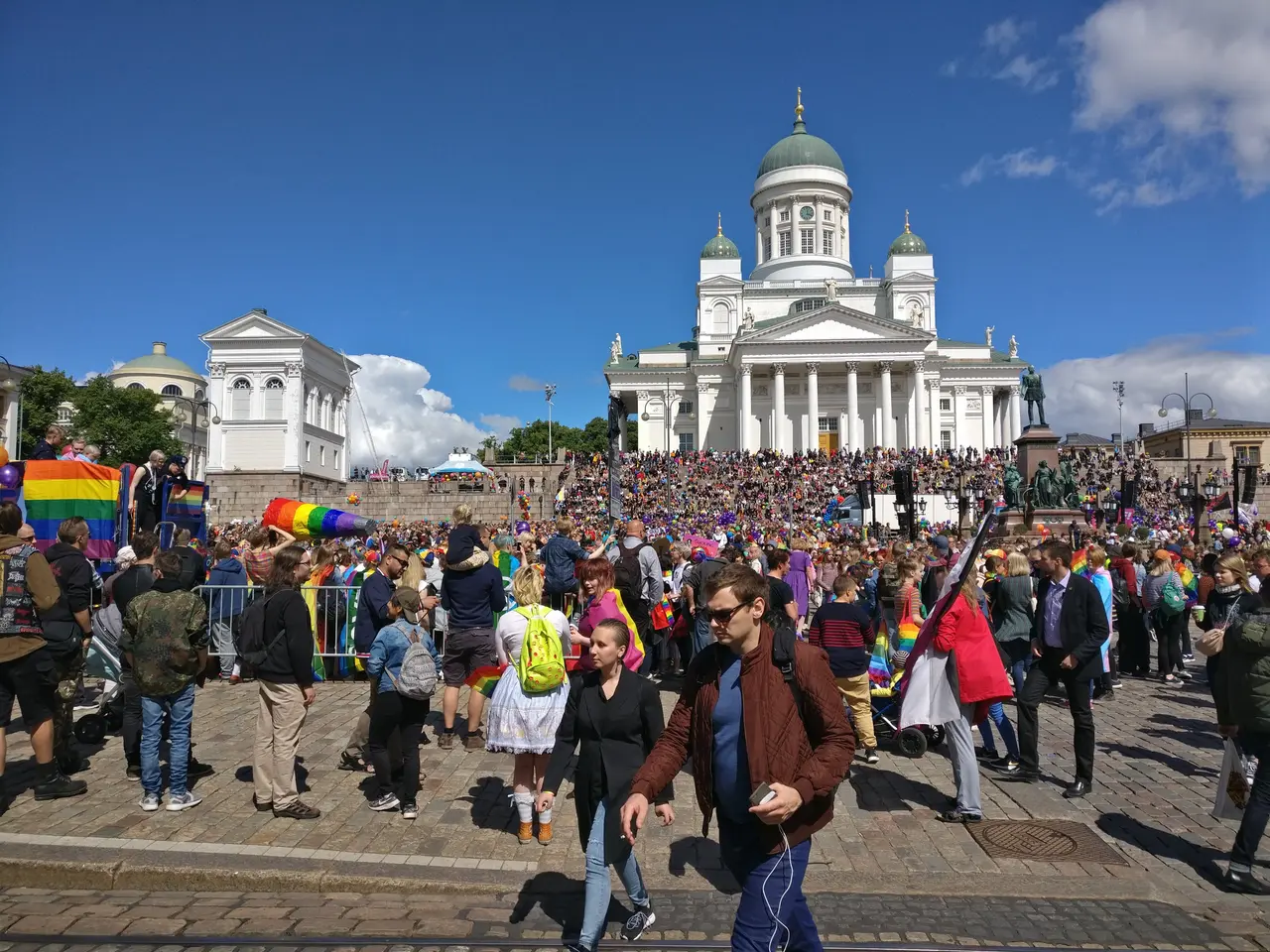 The image shows a large group of people walking down a street in front of a building with windows,...
