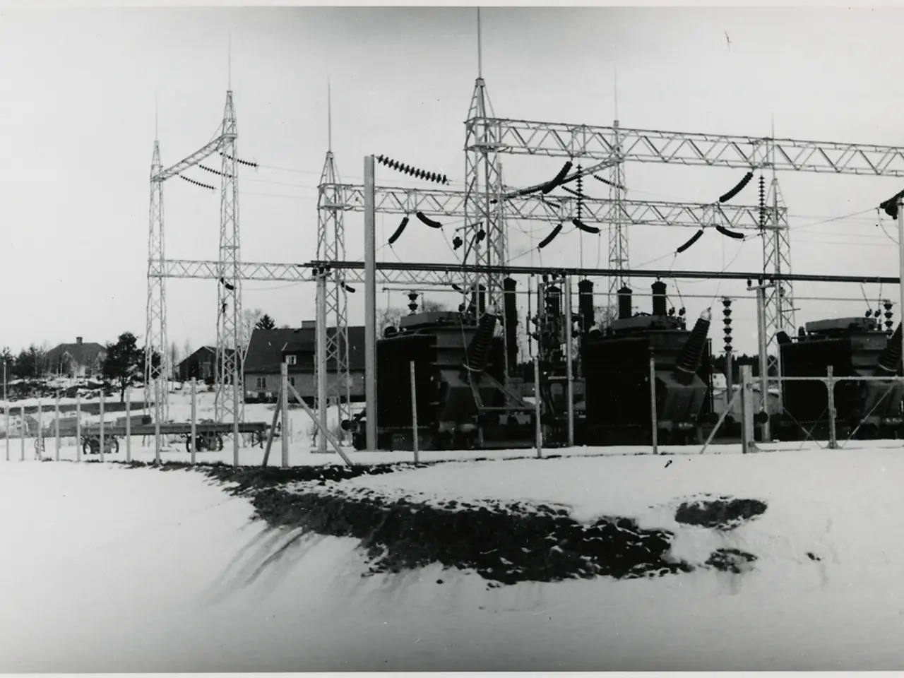 The image shows a black and white photo of an electrical substation in the snow, with transformers,...