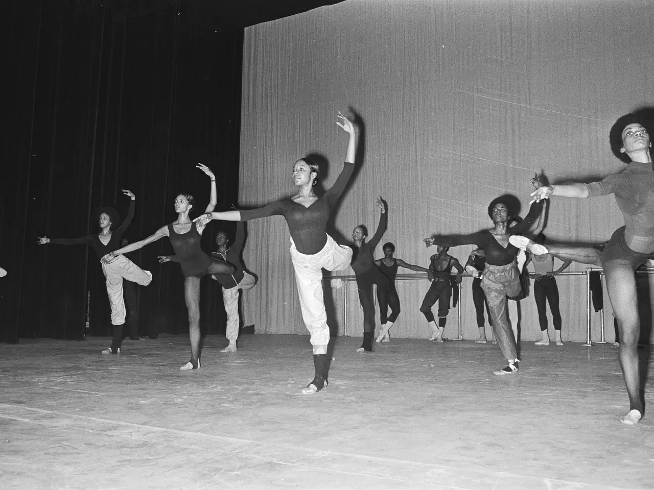 The image shows a group of dancers on a stage with their arms outstretched, wearing black and white...
