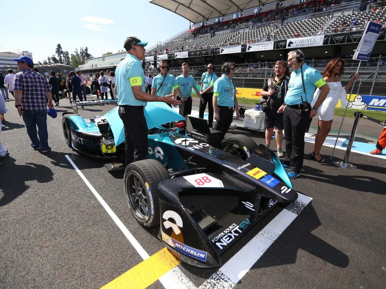 The image shows a group of people standing around a race car on a track, with a stadium in the...